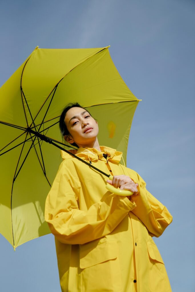 Portrait of an adult wearing a yellow jacket and umbrella under a clear blue sky, captured from a low angle.