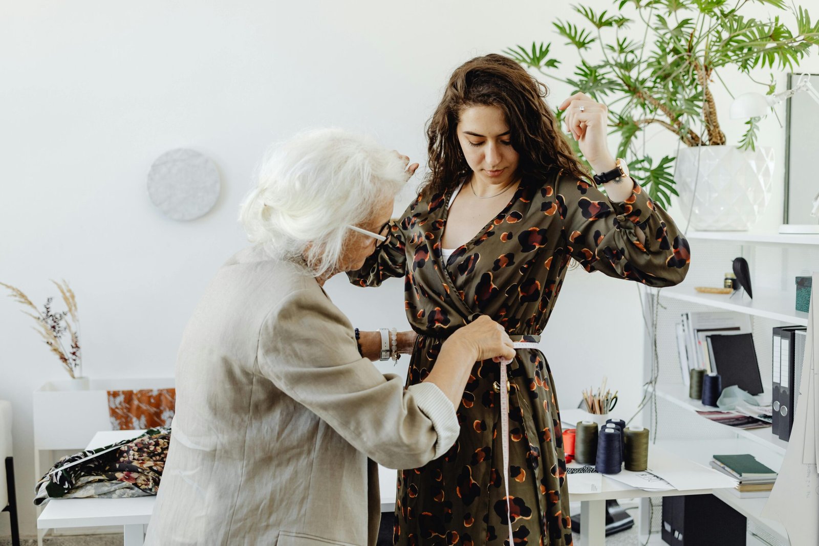 An elderly dressmaker adjusts a young woman's dress indoors, showcasing a personal fashion touch.